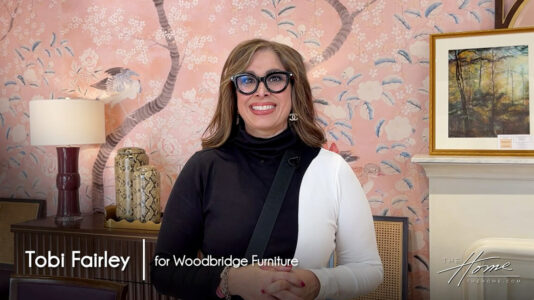 image of interior designer Tobi Farley in black and white dress with big black rimmed glasses, background has pink floral wallpaper . Text: Tobi Fairley for Woodbridge Furniture