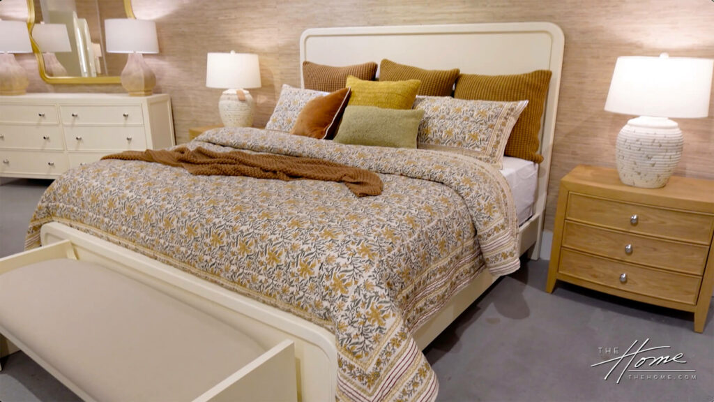 Image: furniture showroom with white rounded headboard and footboard, floral yellow and green patterned bedspread and brown and multicolored pillows, brown throw, wood nightstand and white dresser.
