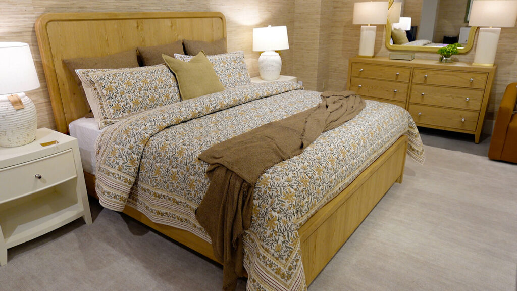 Image: Bassett Newberry Collectionbed with light oak wood headboard and footboard, patterned bedspread with brown throw and home spun pillows, in furniture showroom