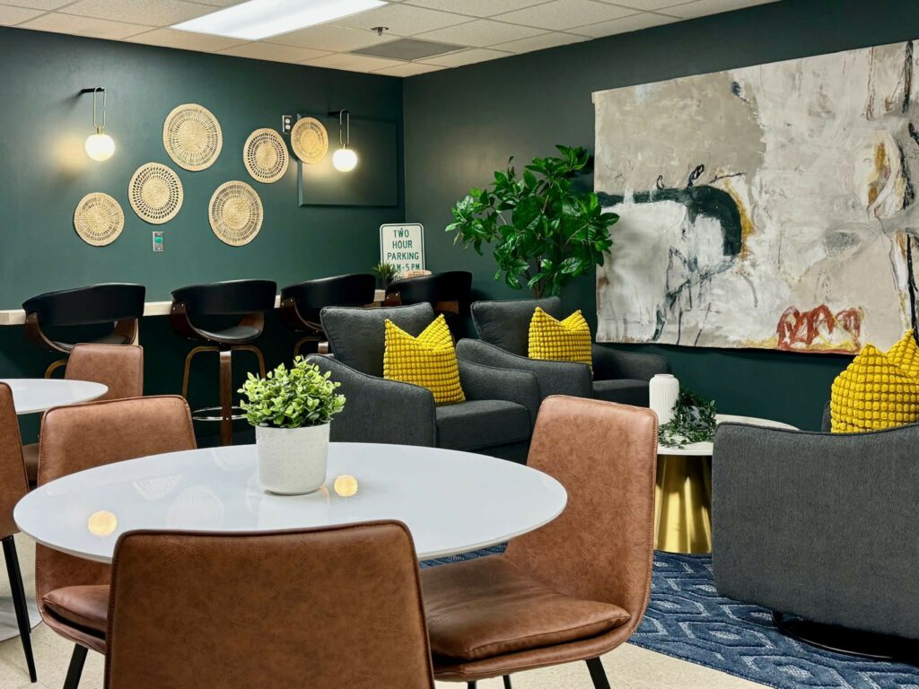 Image: dark green room with abstract art on wall, white round cafe tables and brown chairs, 4 bar stool chairs at bar, gray upholstered chairs with bright yellow pillows, blue diamond pattern rug