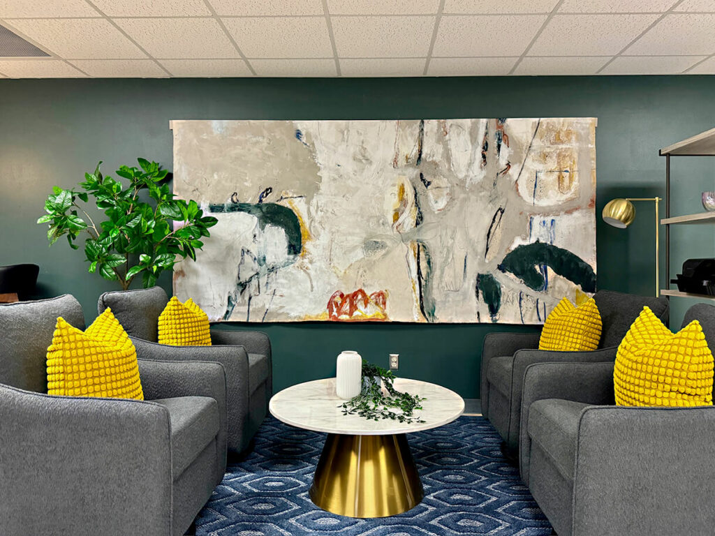 4 gray stuffed upholstered chairs with bright yellow pillows around small marble circular coffee table, blue rug, abstract art on dark green wall