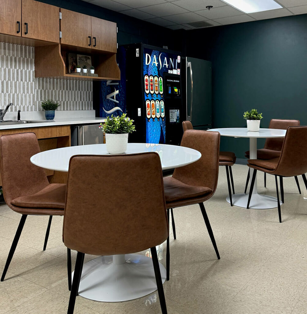 teachers lounge with cafe style round white tables, 3 brown kitchen chairs, backsplash on kitchen wall with brown cabinets, dark green wall with vending machine