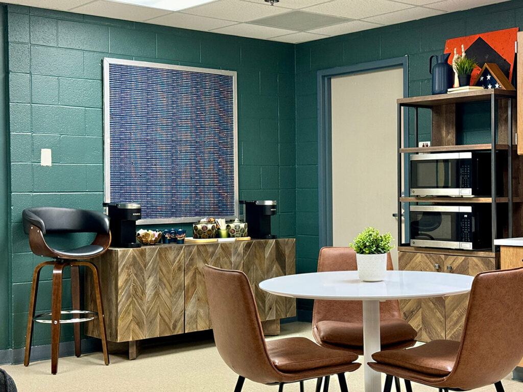dark green painted cinderblock wall with 2 microwaves in modern wood bureau containing zigzag pattern inlay, black modern barstool chair, abstract green and blue artwork, wood shelving, cafe table and door