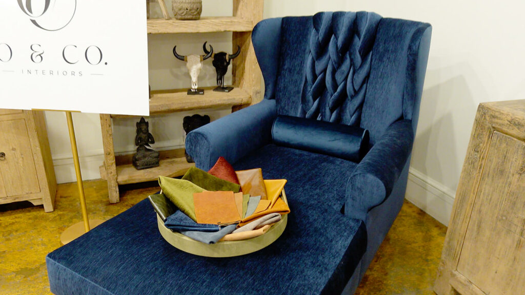 Image: O & Company Victoria Lane sapphire blue velvet upholstered chair with braid on seat back, bowl with leather and fabric samples in showroom with wood floor