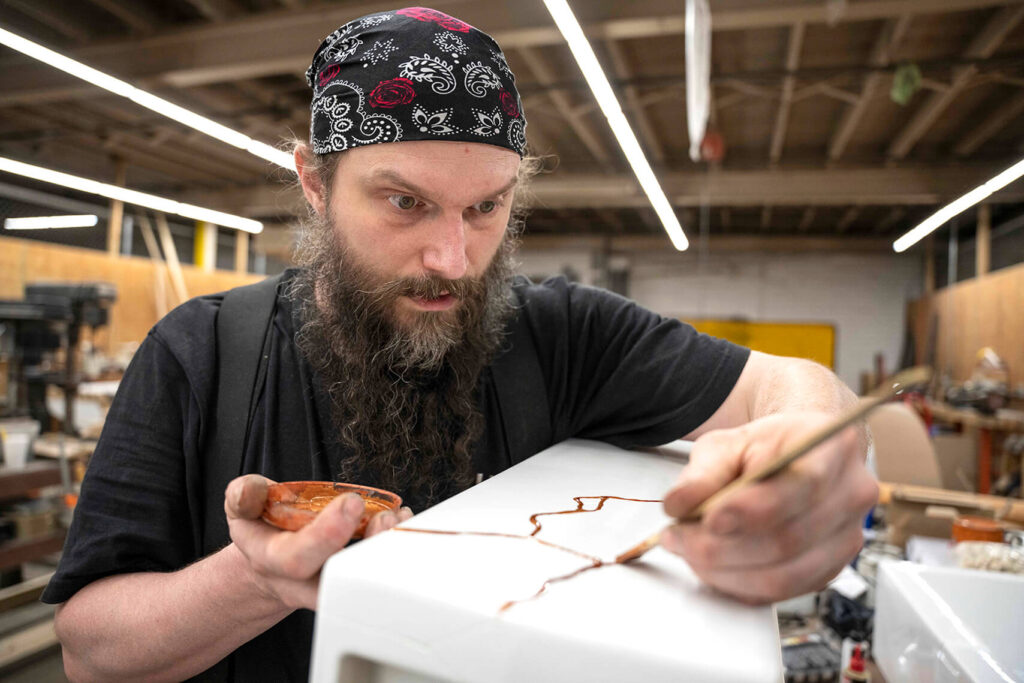 Image: artist Trevor Waring, man with a beard and head bandana, working with copper on Thompson Traders Kintsugi Collection fireclay sink in warehouse