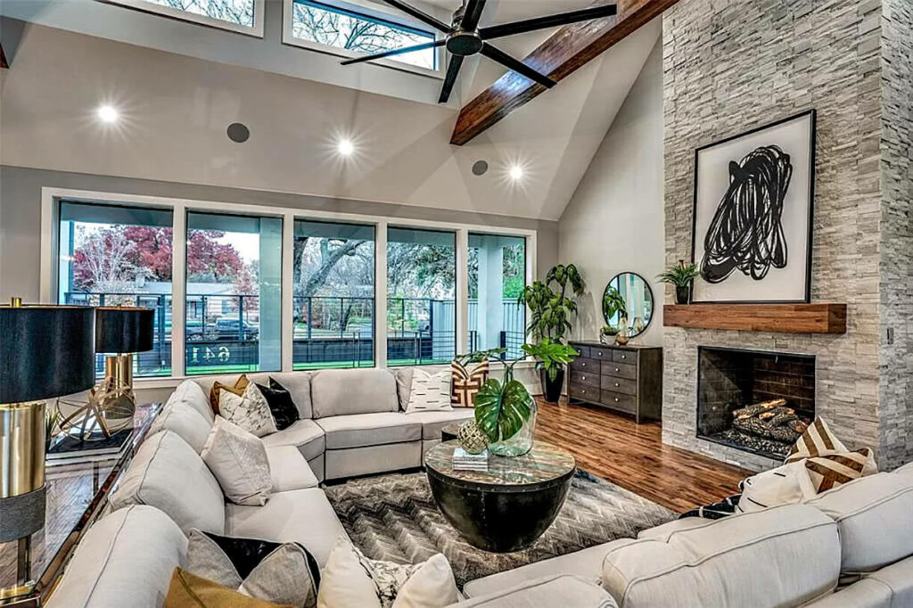 Image: Great room with vaulted wood beam ceiling, u-shape sectional sofa in beige, fireplace with wood mantle,  black and brass lamps, skylights