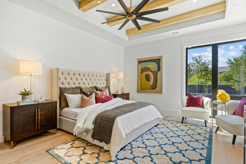 Image: white wall bedroom, vaulted ceiling with wood beams & fan, upholstered headboard bed, throw pillows, white and blue design carpet, cozy atmosphere