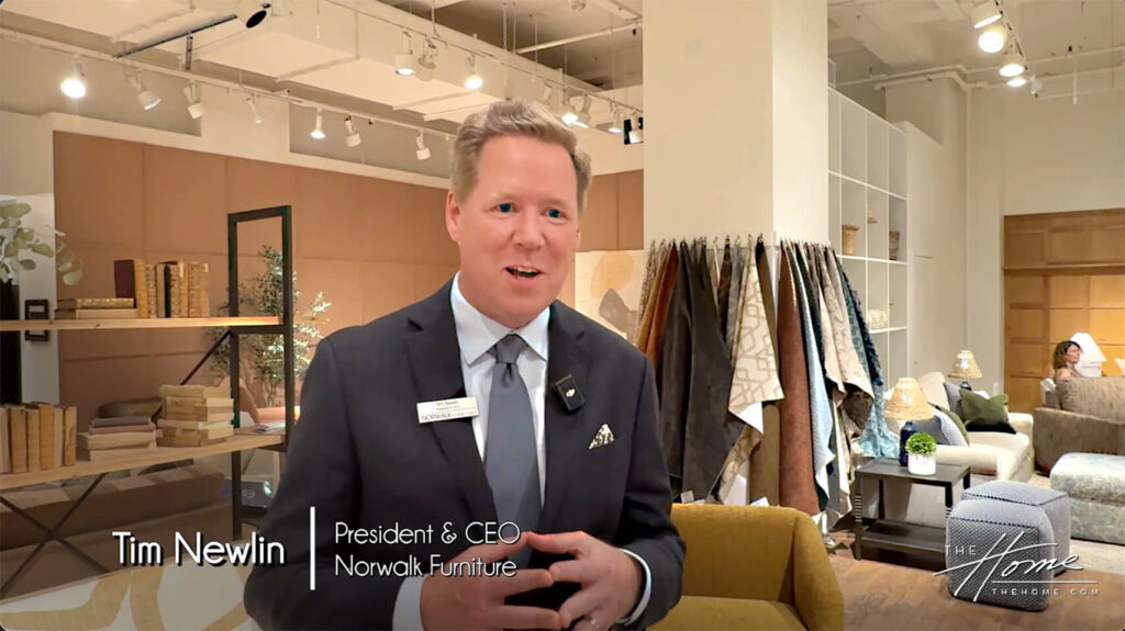 Image: Blonde man in dark suit in Norwalk Furniture showroom with fabric samples hanging and brown chair, bookshelf and lights